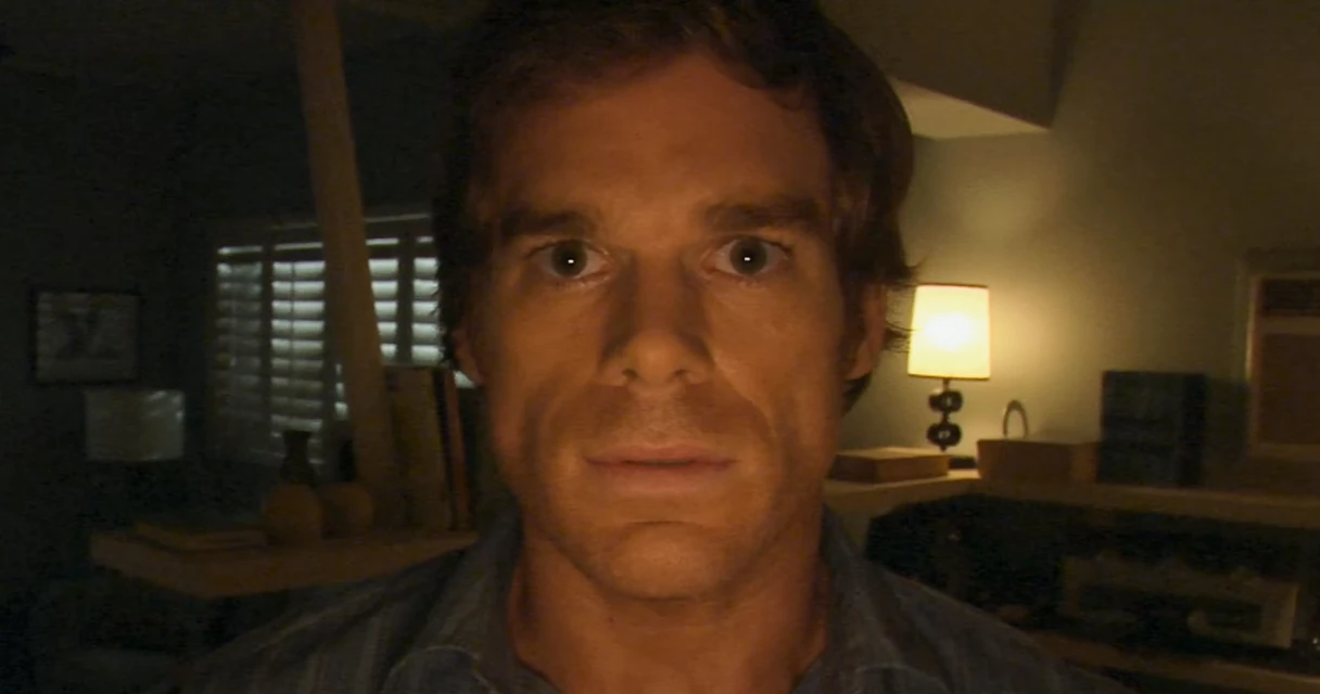 Dexter Morgan from Dexter sitting in a dimly lit room with a lamp behind him, staring blankly into the camera, no text